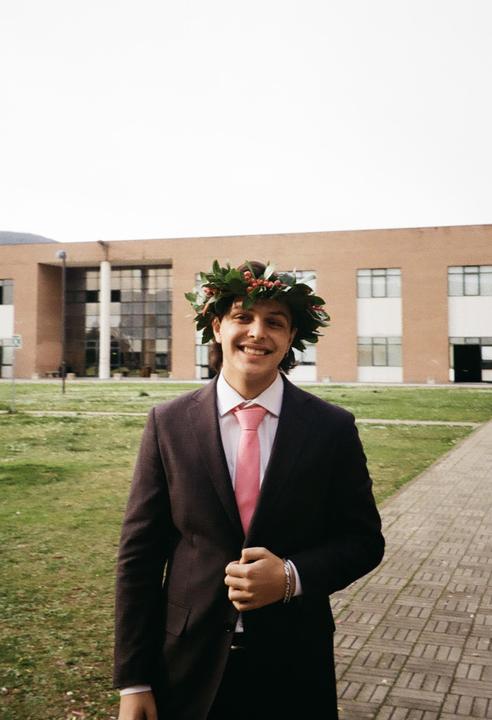 A man wearing a suit and a wreath of leaves and berries smiles at the camera in front of a building.