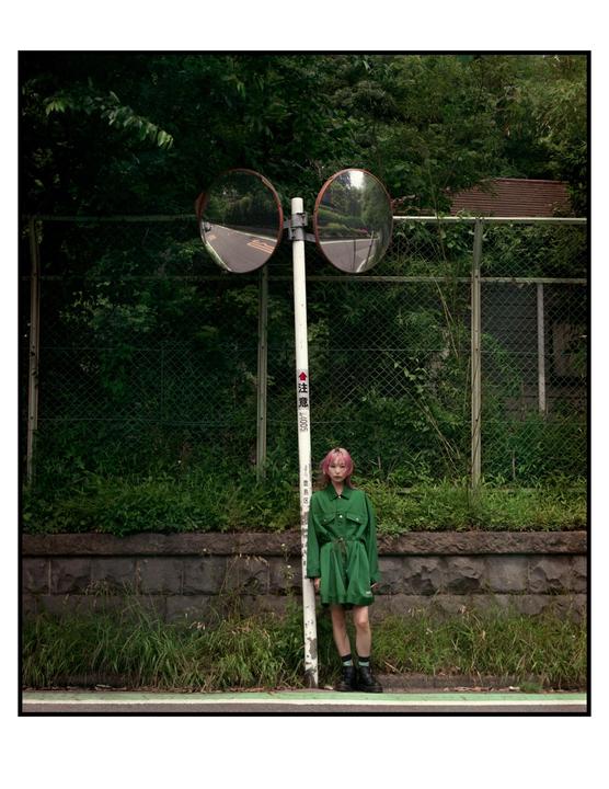 A woman with pink hair stands next to a pole with two convex mirrors on a street.