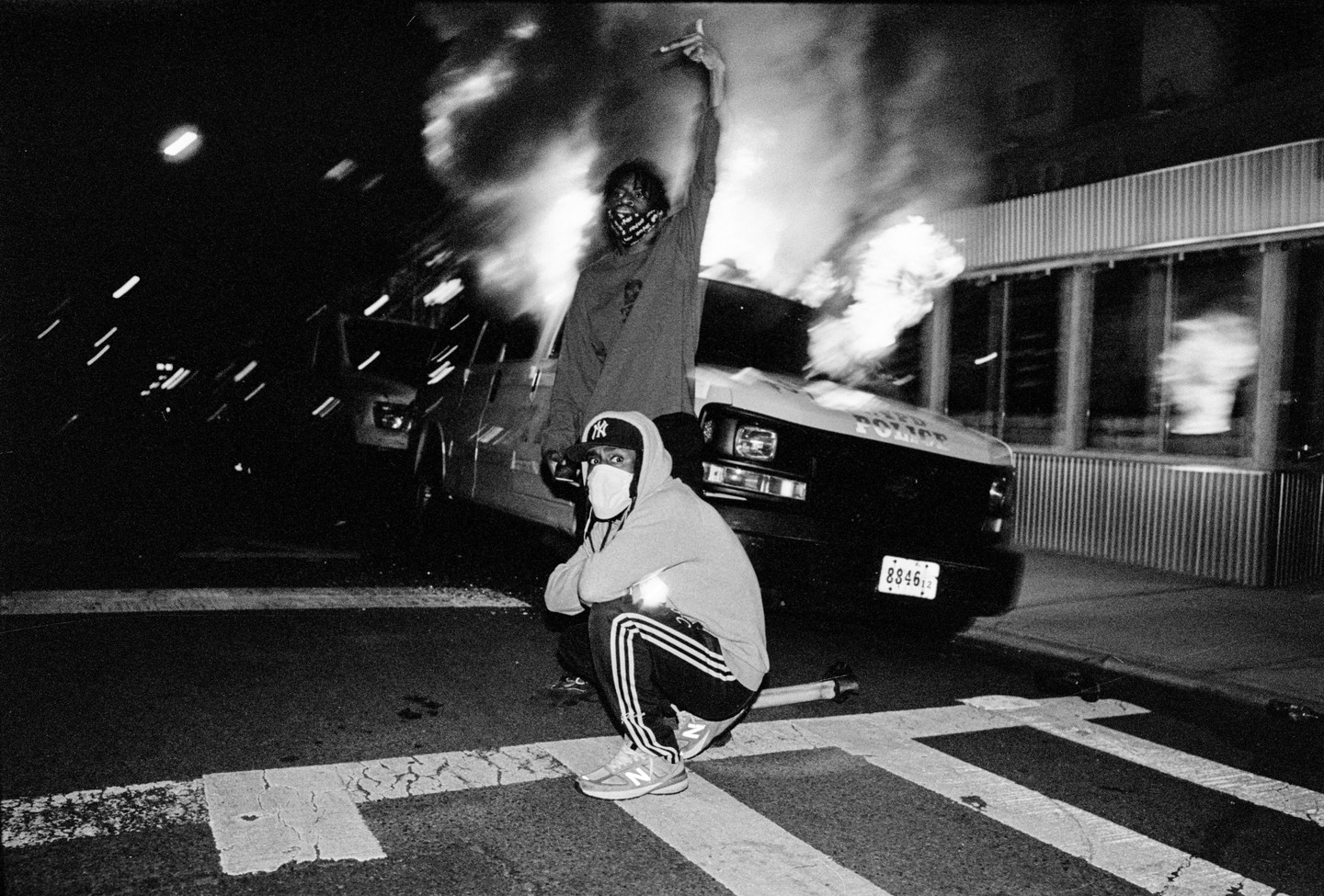 Two men stand in front of a burning police vehicle at night.