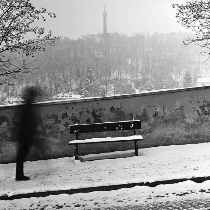 A blurry figure walks past a snow-covered bench with a distant tower visible above a wooded hill.
