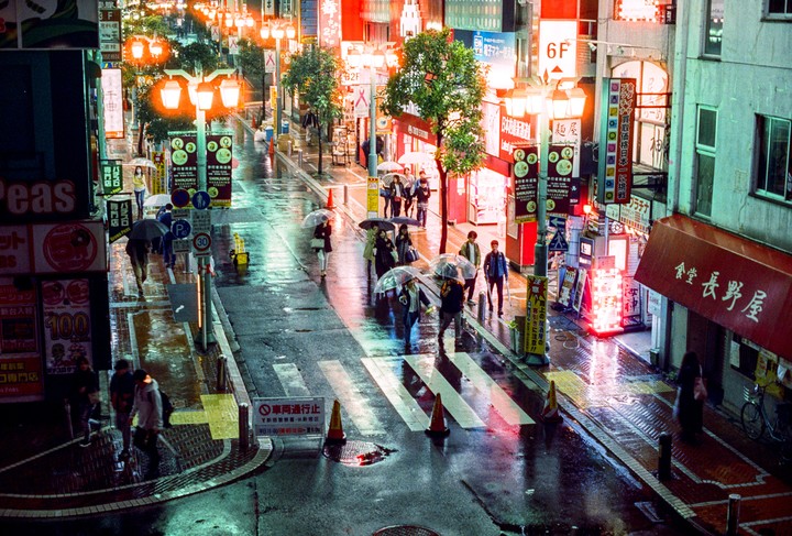 People walk with umbrellas on a wet street lined with neon signs and lanterns at night in Shinjuku.
