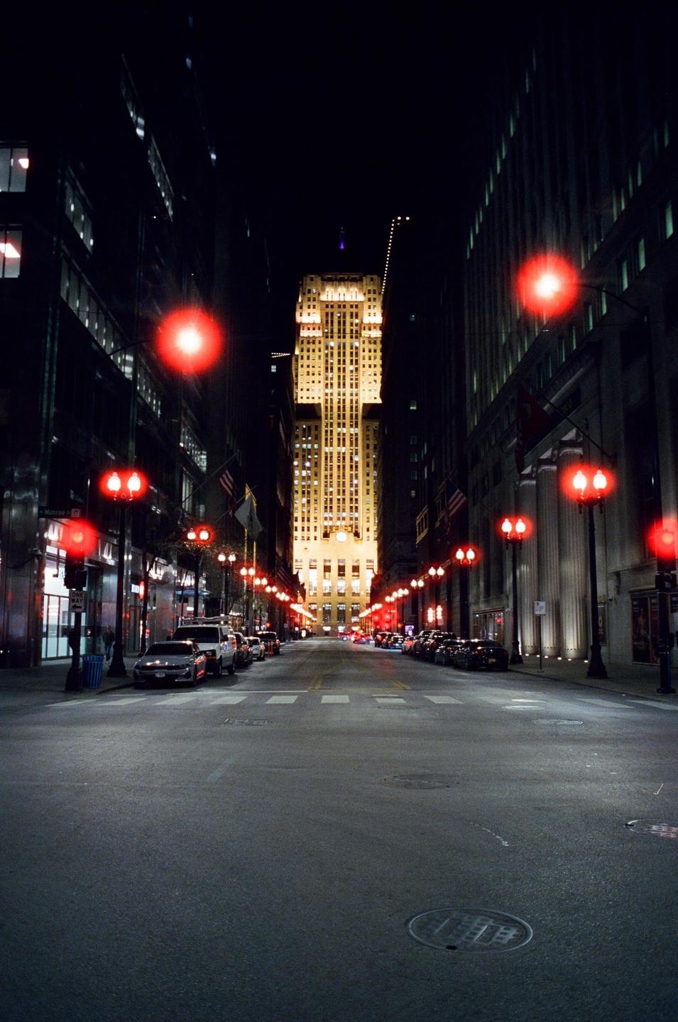 A street at night is lined with parked cars and red streetlights, leading to a brightly lit skyscraper in the distance.