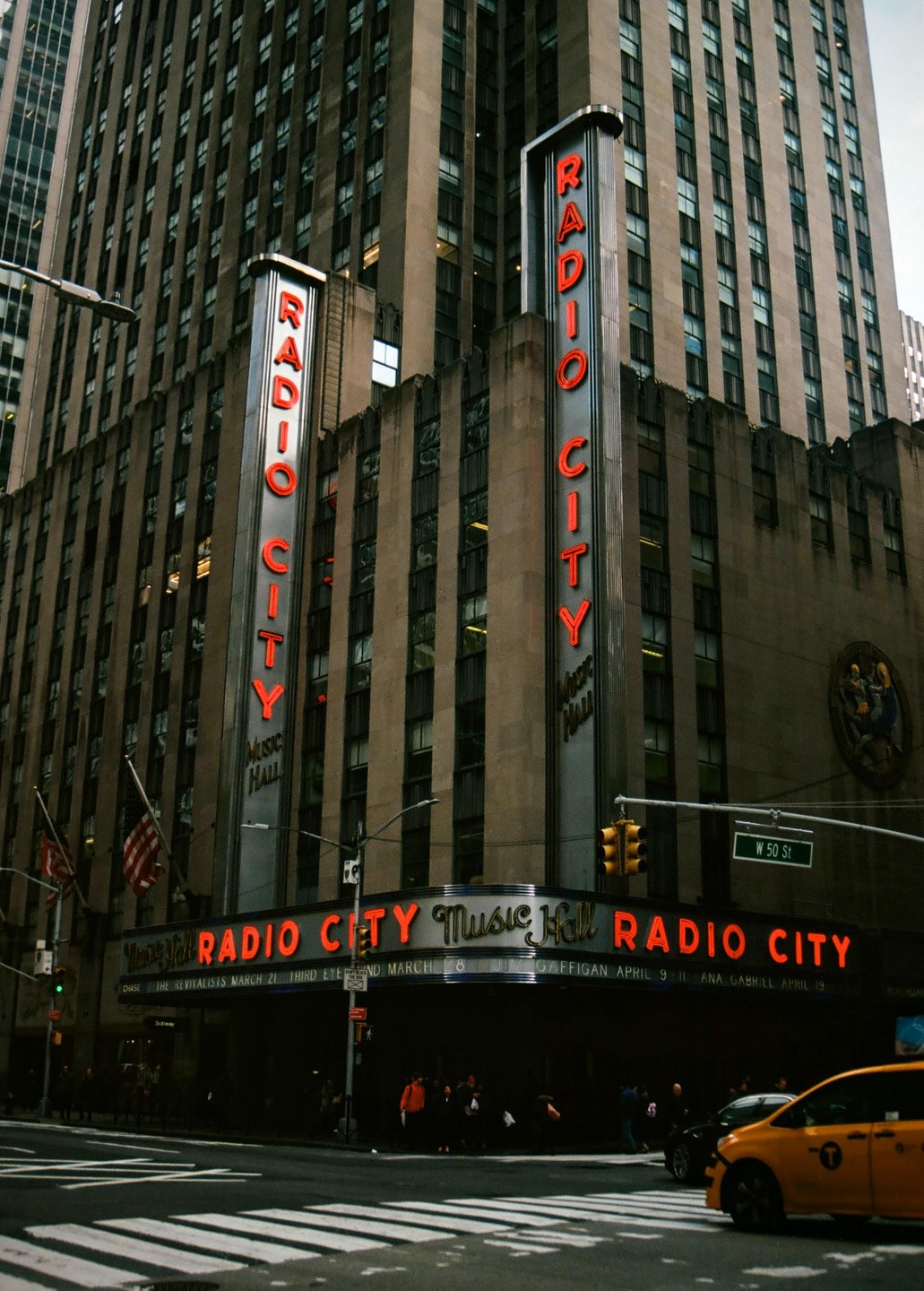 The Radio City Music Hall building with its neon signs is visible from a street corner with a yellow taxi cab and pedestrians.