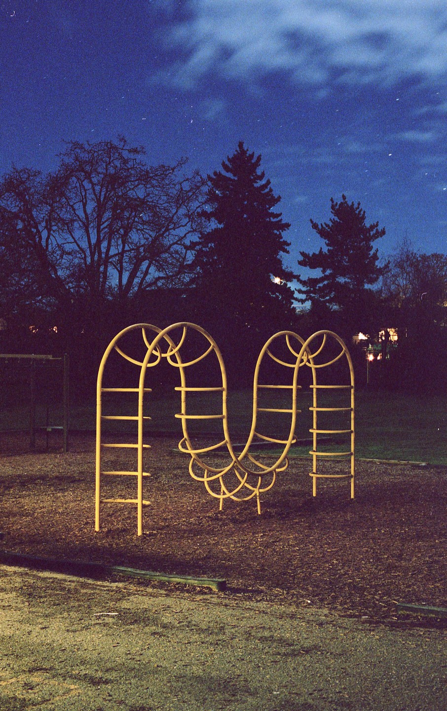 A yellow climbing frame stands in a playground at night, with trees and a dark sky in the background.