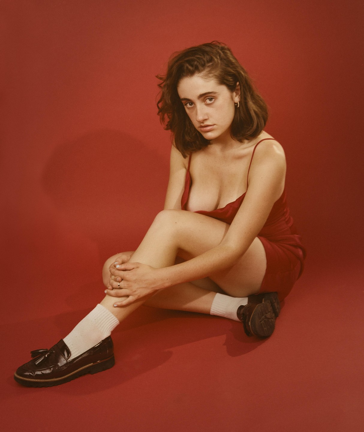A woman in a red dress and loafers sits on a red surface.