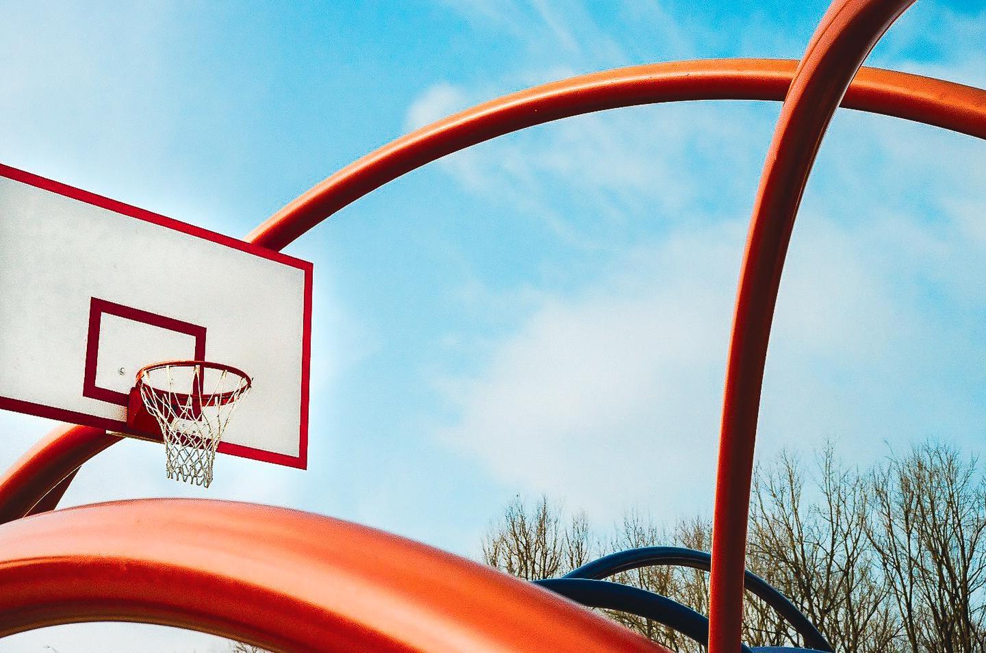 A basketball hoop and backboard are visible against a blue sky with abstract orange and blue structures in the foreground.