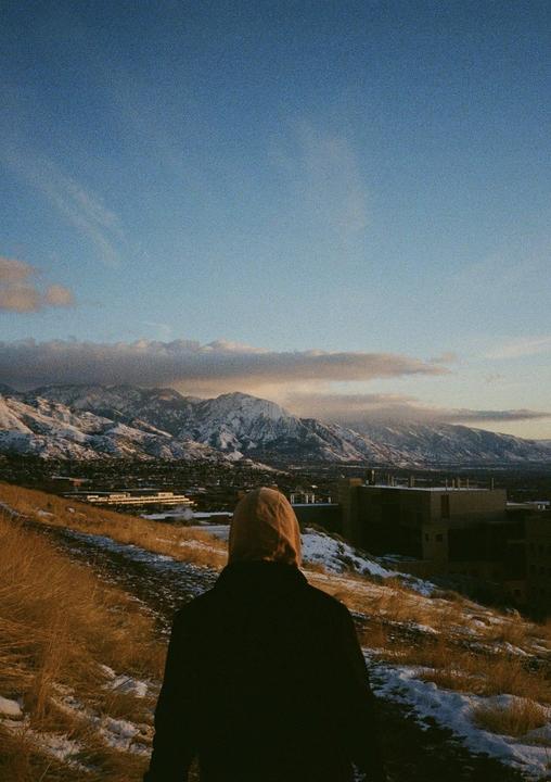 A person in a hooded jacket stands on a snowy hill overlooking a town and snow-capped mountains under a blue sky.