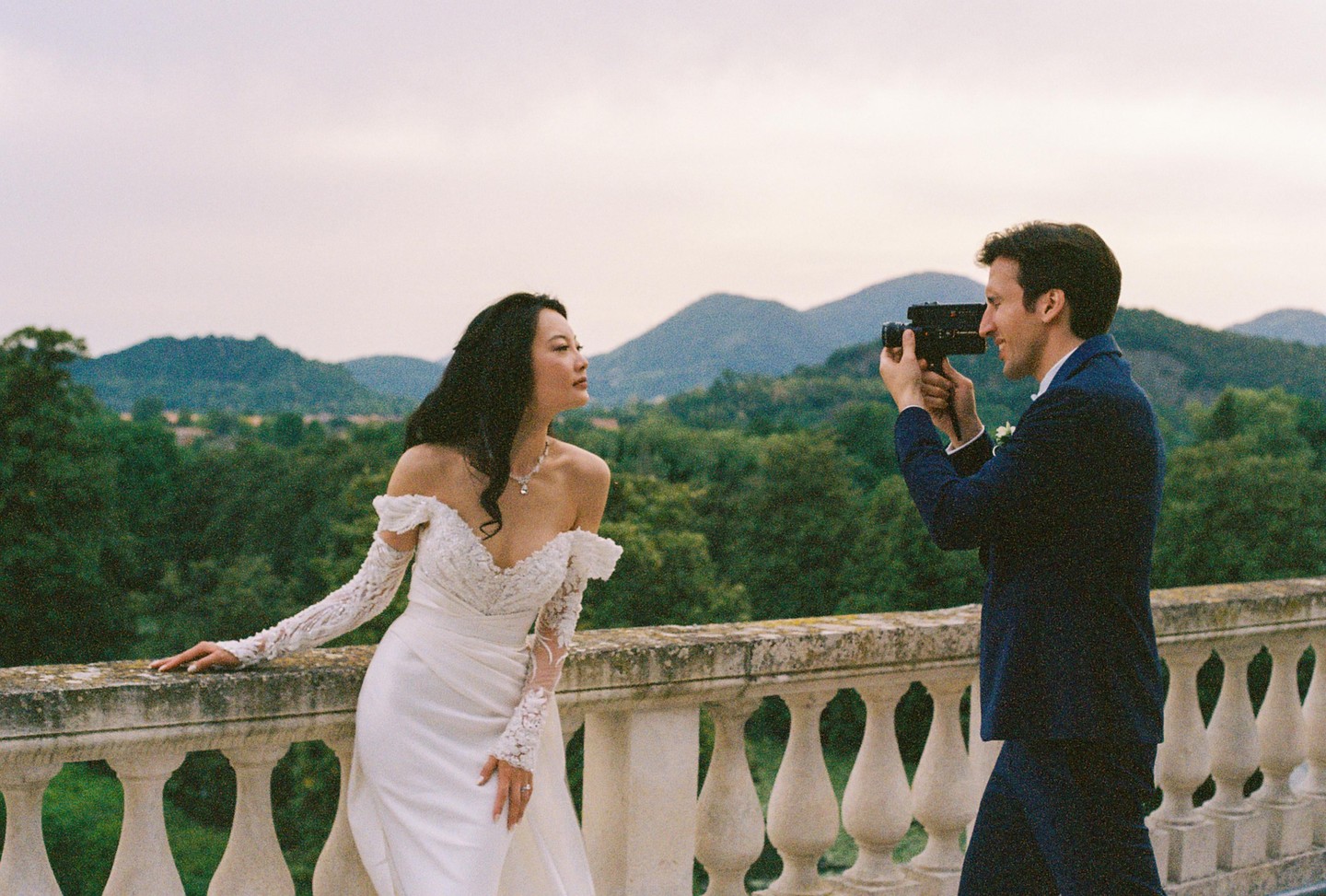 A woman in a wedding dress poses on a balcony while a man films her with a video camera, with mountains and trees in the background.