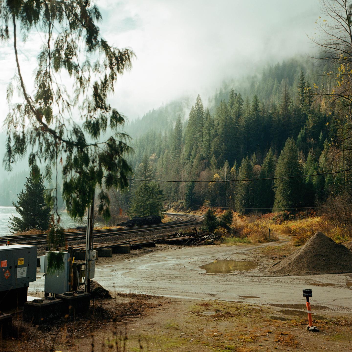 A winding railroad track runs alongside a body of water with a misty, forested mountain in the background.