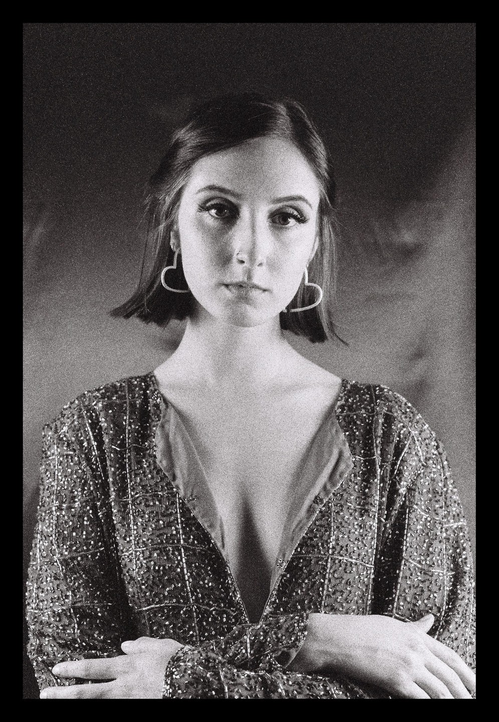 A woman with short hair and heart-shaped earrings wears a sequined top and looks directly at the viewer.