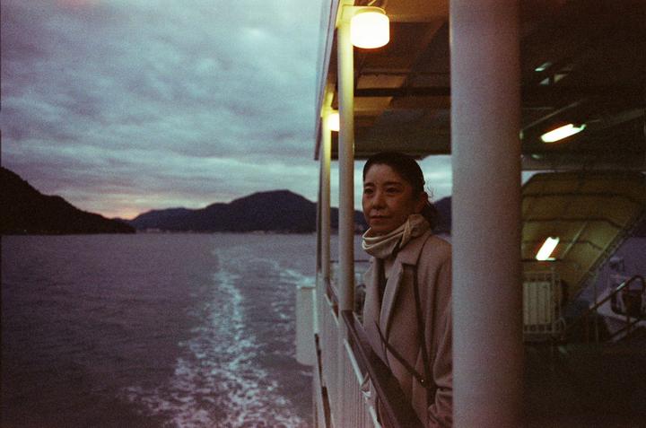 A woman stands on a boat deck at dusk, looking out at the water and mountains.