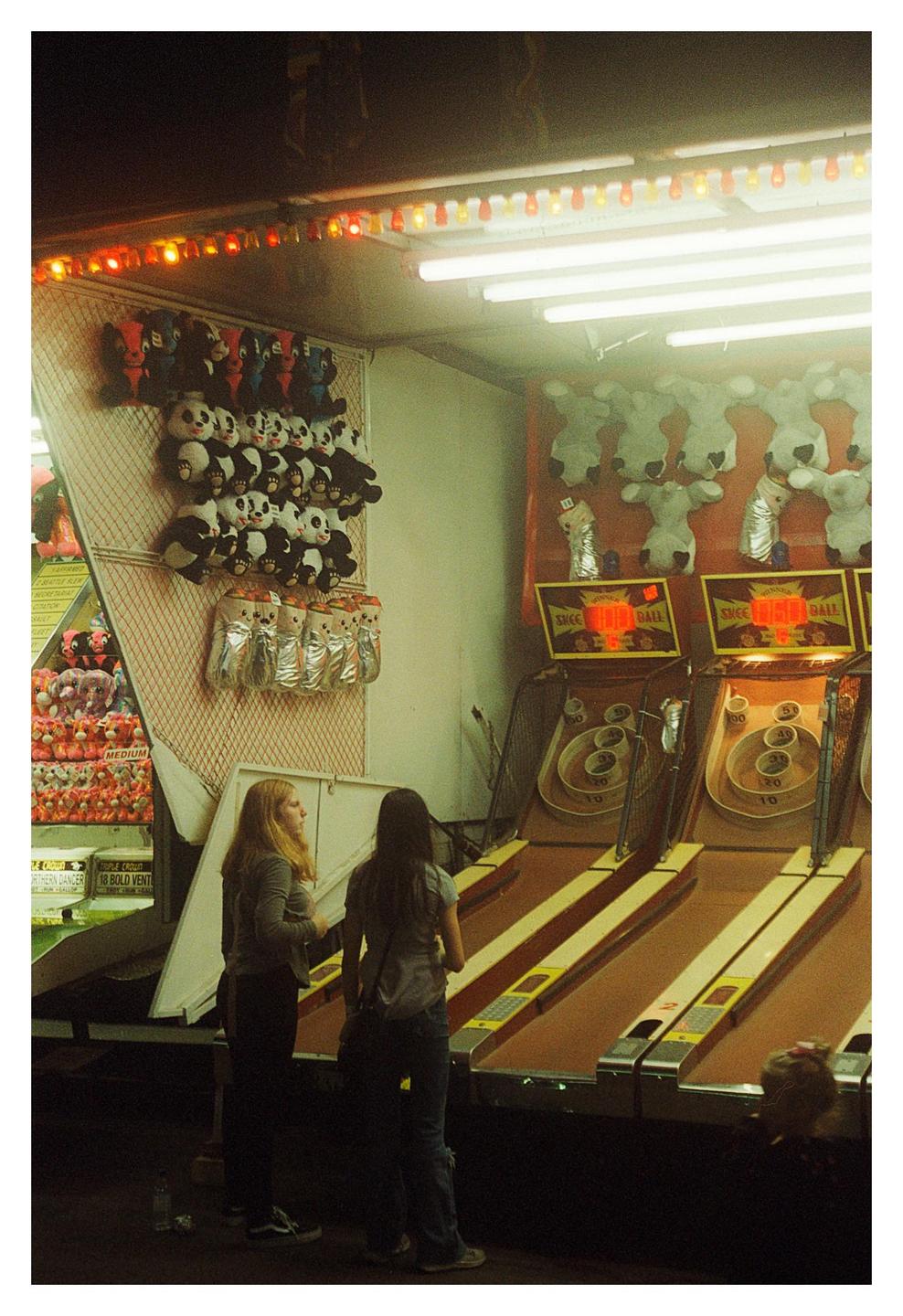 Two women stand in front of a carnival game with stuffed animals hanging on the wall.