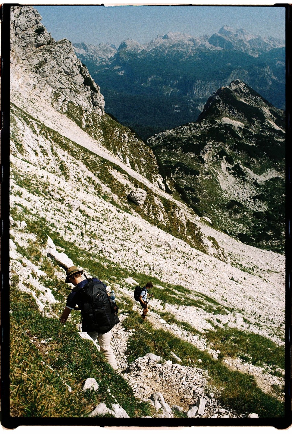 Two men hike up a steep, rocky mountain path with a vast mountain range in the background.