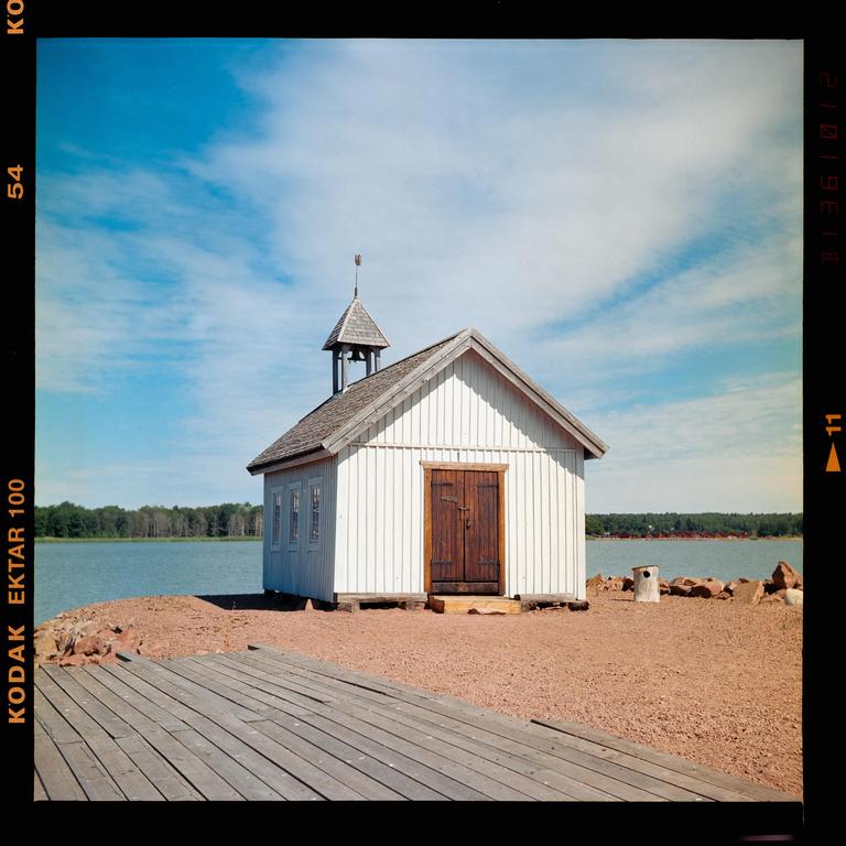 A small white chapel with a bell tower stands on a rocky shore next to a body of water, with a wooden deck in the foreground.