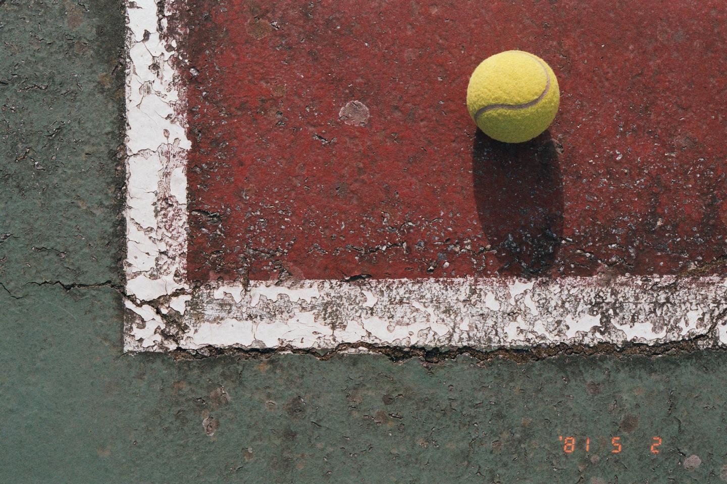 A tennis ball rests on the red court next to a white line, with a green court visible in the bottom left.