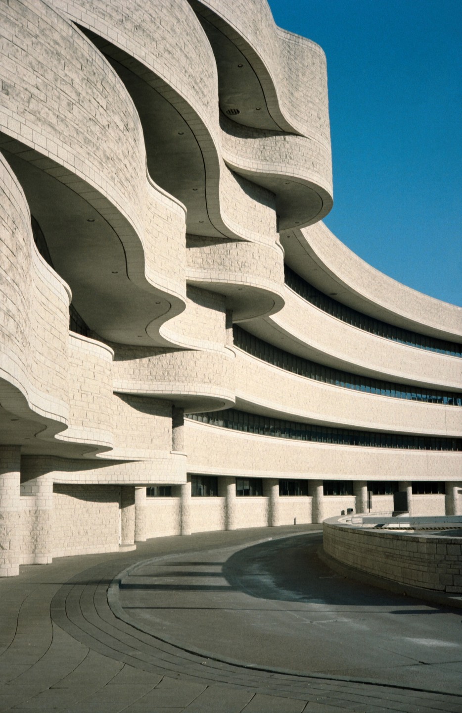 A modern building with curved, textured walls and balconies is shown under a clear blue sky.