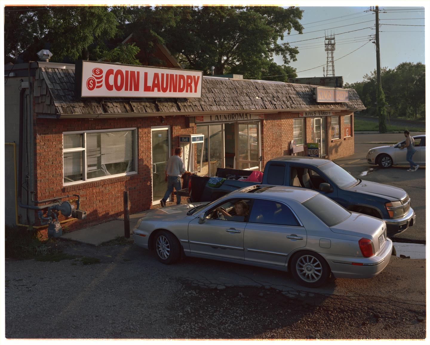 A man walks into a coin laundry building with cars parked outside.