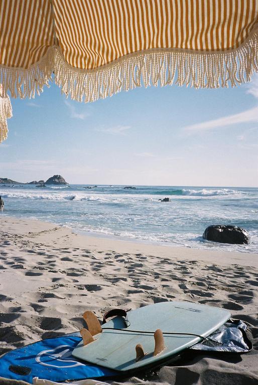 A surfboard rests on a sandy beach with an umbrella overhead and ocean waves in the background.