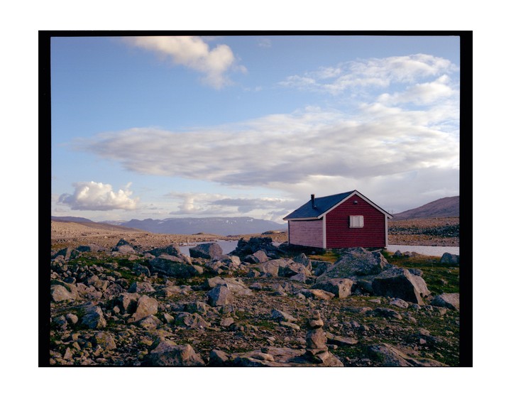 A small red and white cabin sits among rocks and sparse vegetation with mountains and a body of water in the background under a cloudy sky.