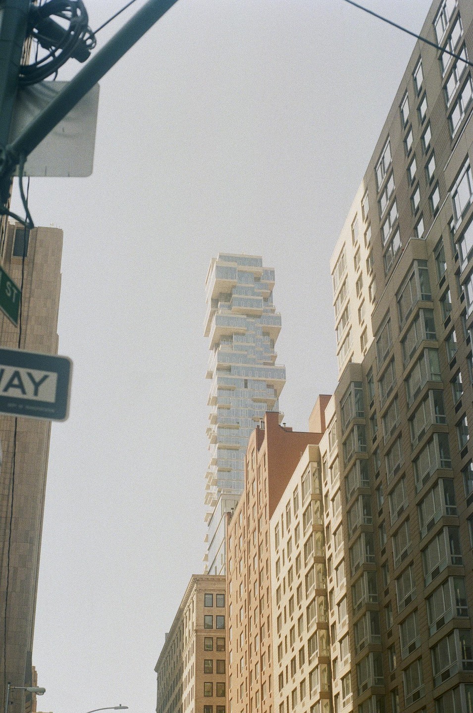 A tall, modern building with many balconies rises above other buildings and a street sign.
