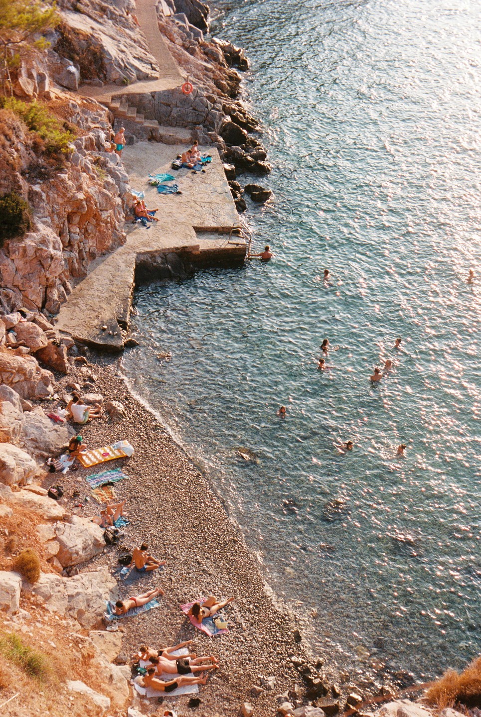 A high-angle view shows people swimming and sunbathing on a rocky beach with a concrete pier.