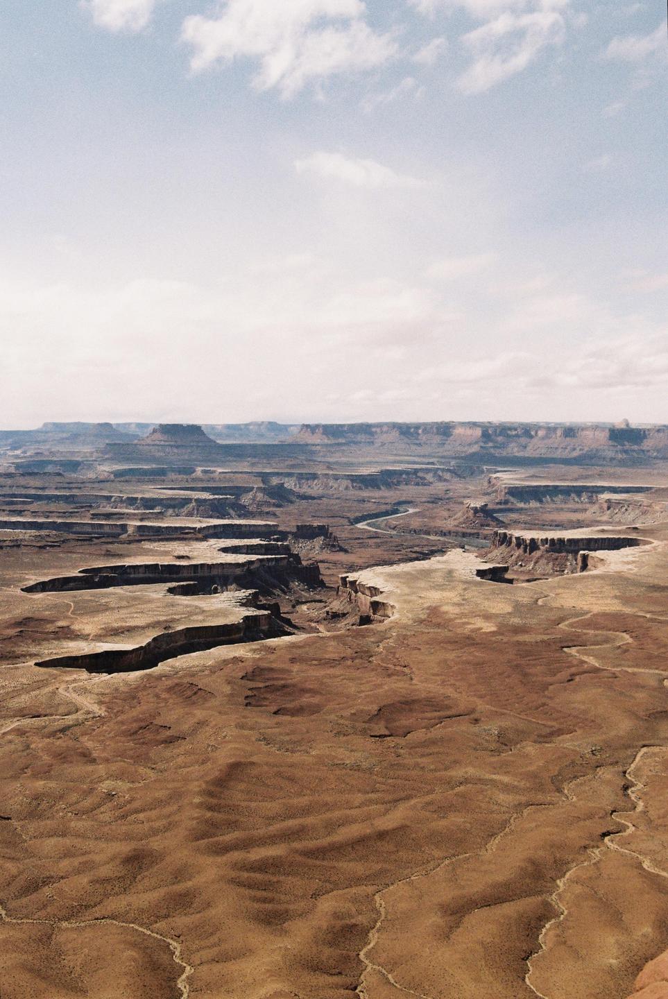 A vast canyon landscape with winding rivers and flat-topped mesas stretches under a partly cloudy sky.
