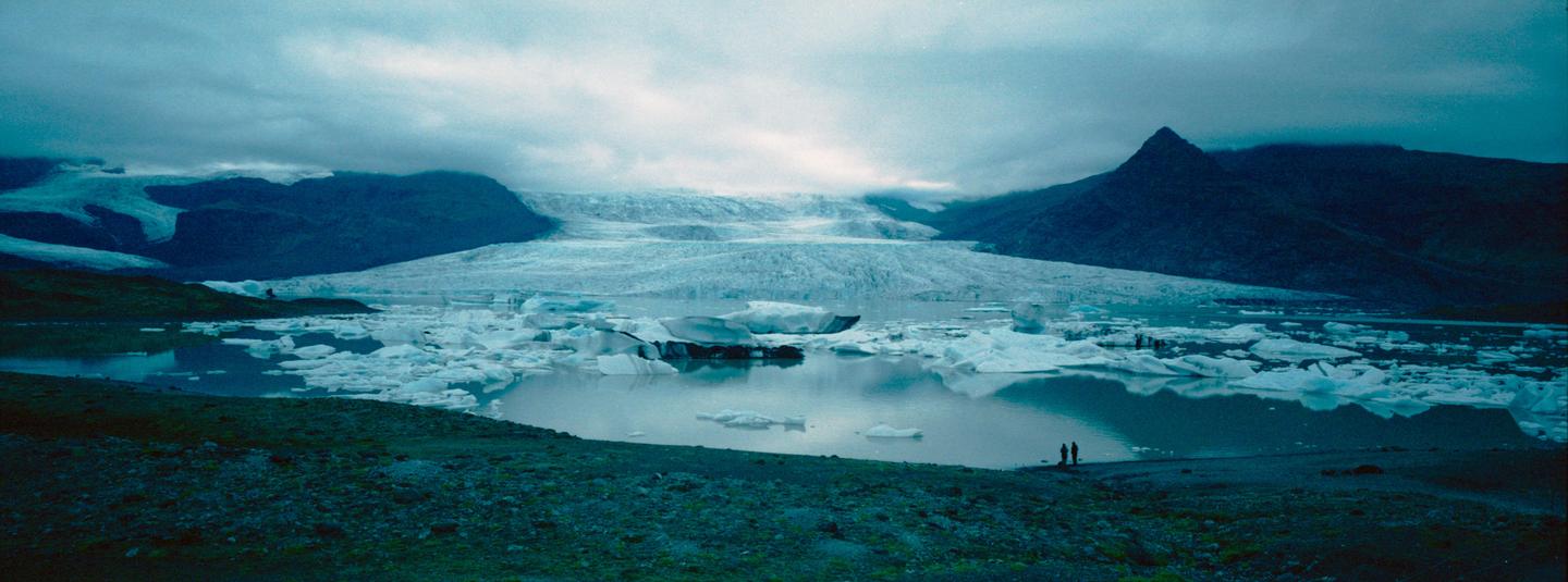 A glacial lagoon with icebergs floating in the water is surrounded by mountains under a cloudy sky.