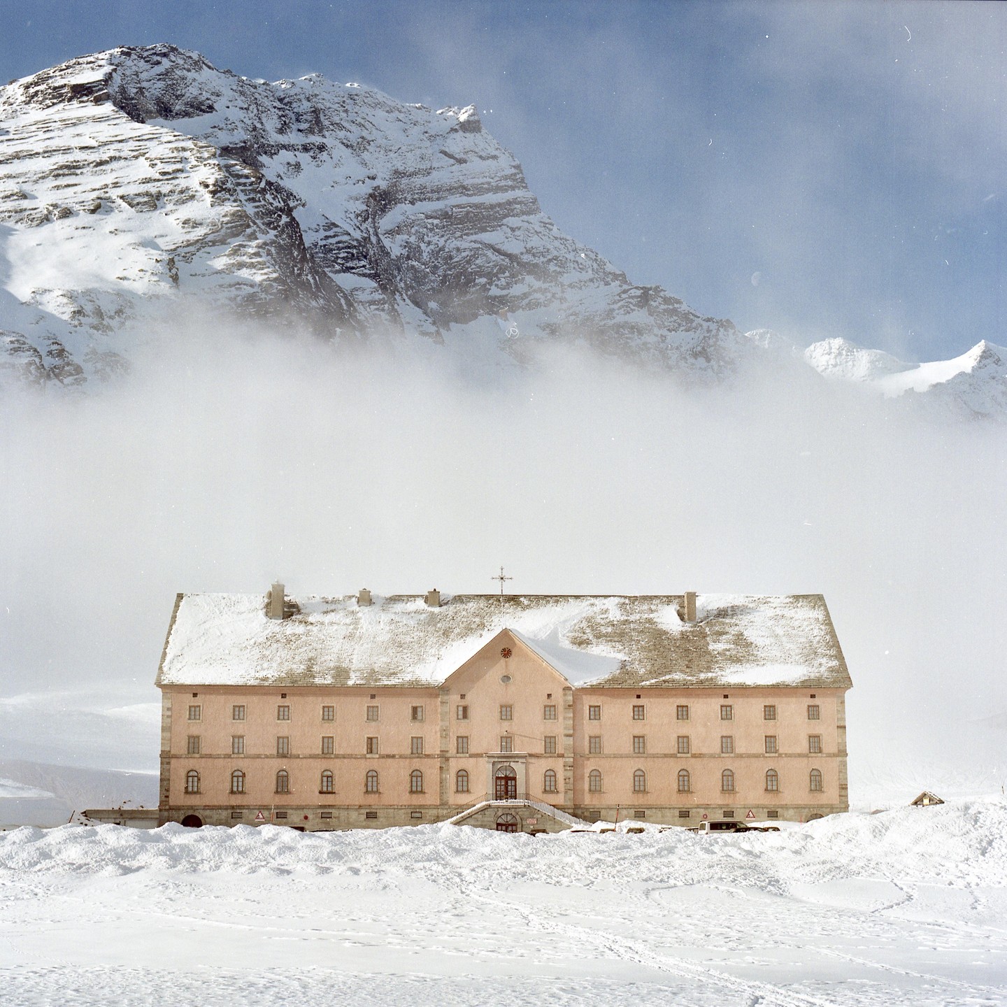 A large, light pink building with a snow-covered roof stands in a snowy landscape with a massive, cloud-shrouded mountain in the background.
