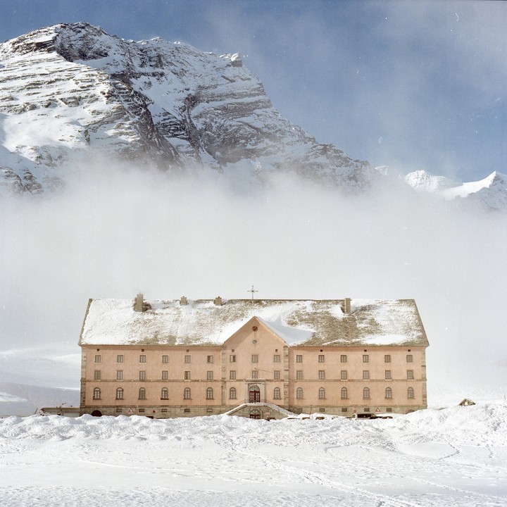 A large, light pink building with a snow-covered roof stands in a snowy landscape with a massive, cloud-shrouded mountain in the background.