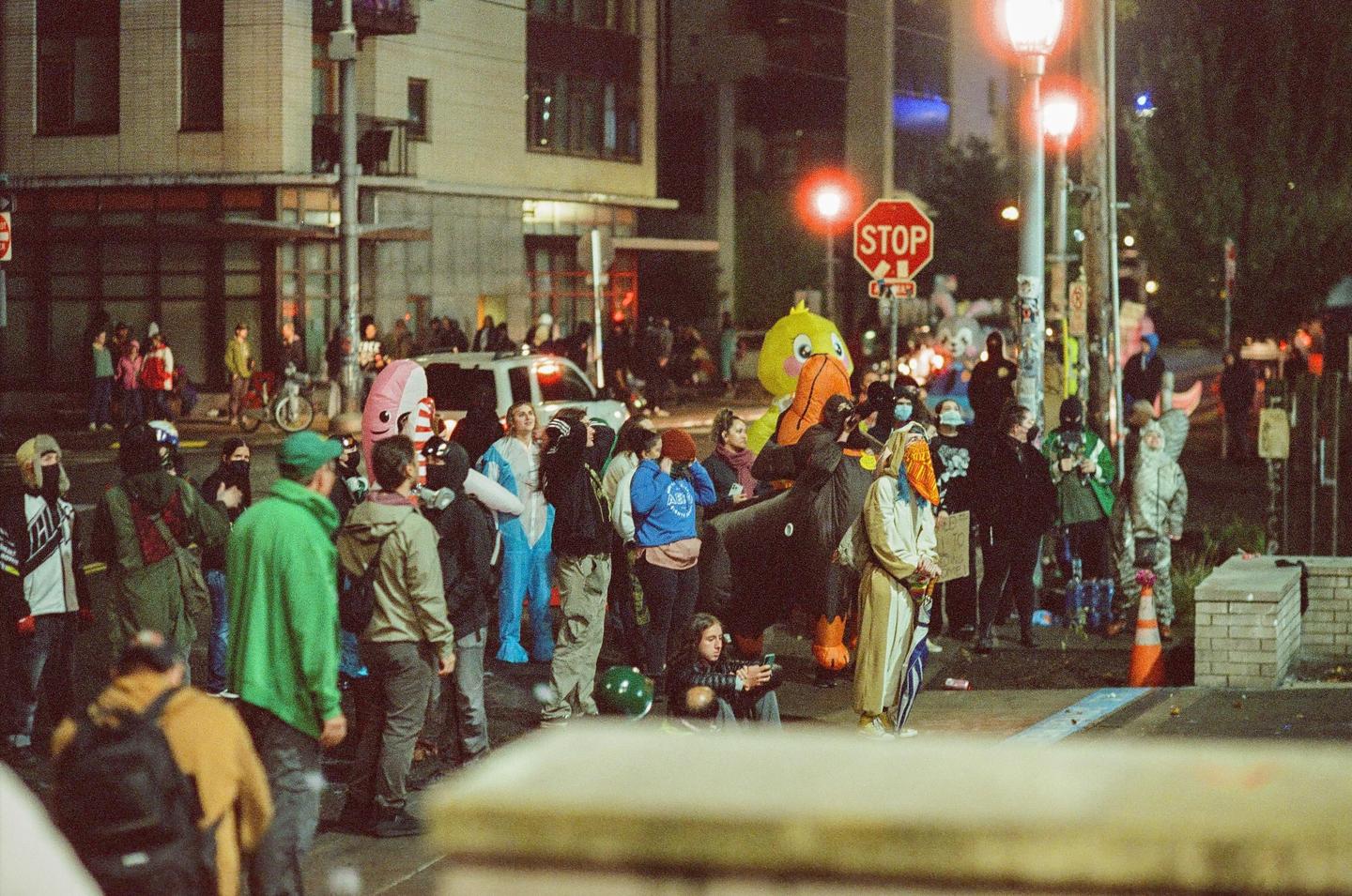 A crowd of people in costumes and masks gather on a city street at night, with a stop sign and buildings in the background.