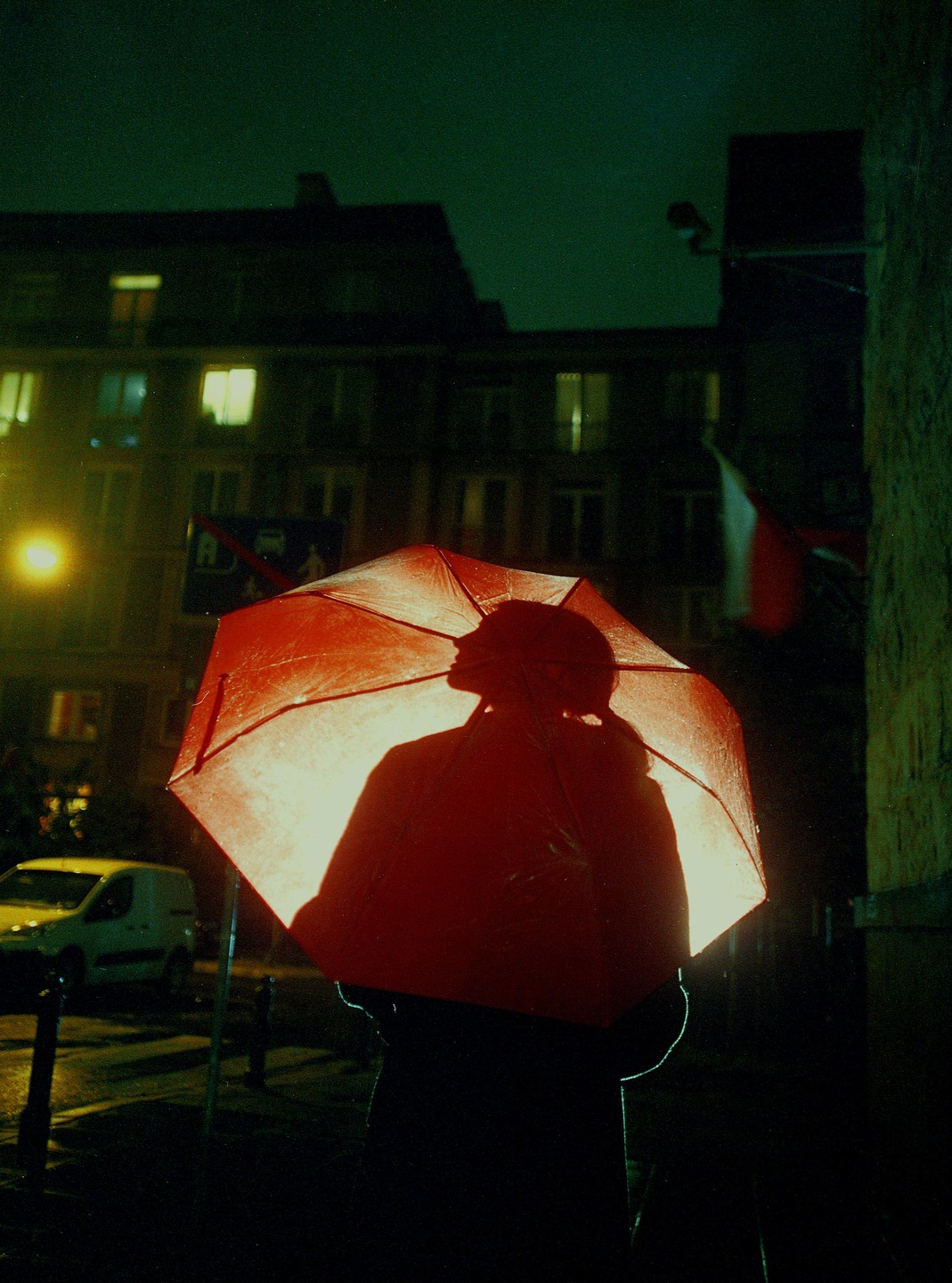 A person holding a red umbrella is silhouetted against a bright light on a dark street at night.