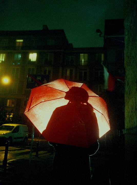 A person holding a red umbrella is silhouetted against a bright light on a dark street at night.
