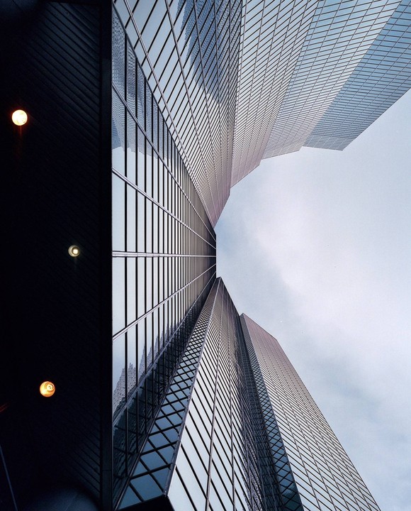 A low-angle view shows the glass facade of a skyscraper reflecting the sky and another building.