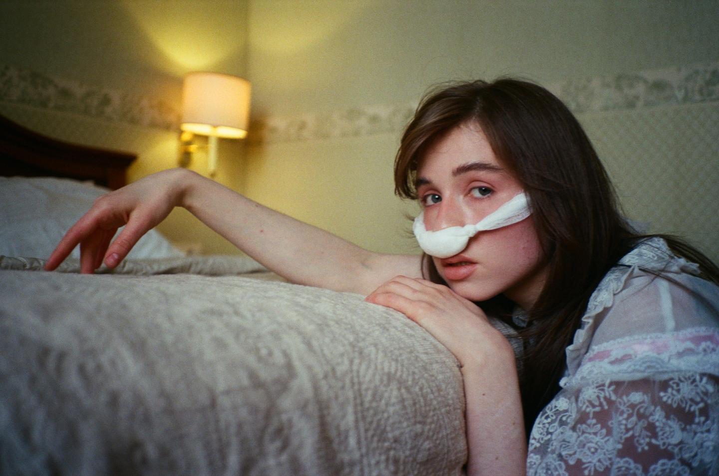 A woman with a bandage on her nose is resting on a bed next to a lamp.