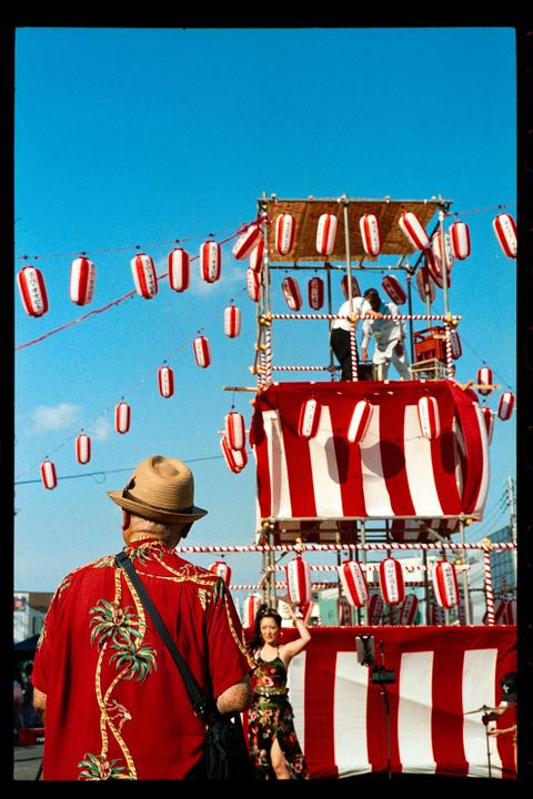 A man in a red Hawaiian shirt and a straw hat stands with his back to the camera, looking at a festival float decorated with red and white lanterns and fabric.