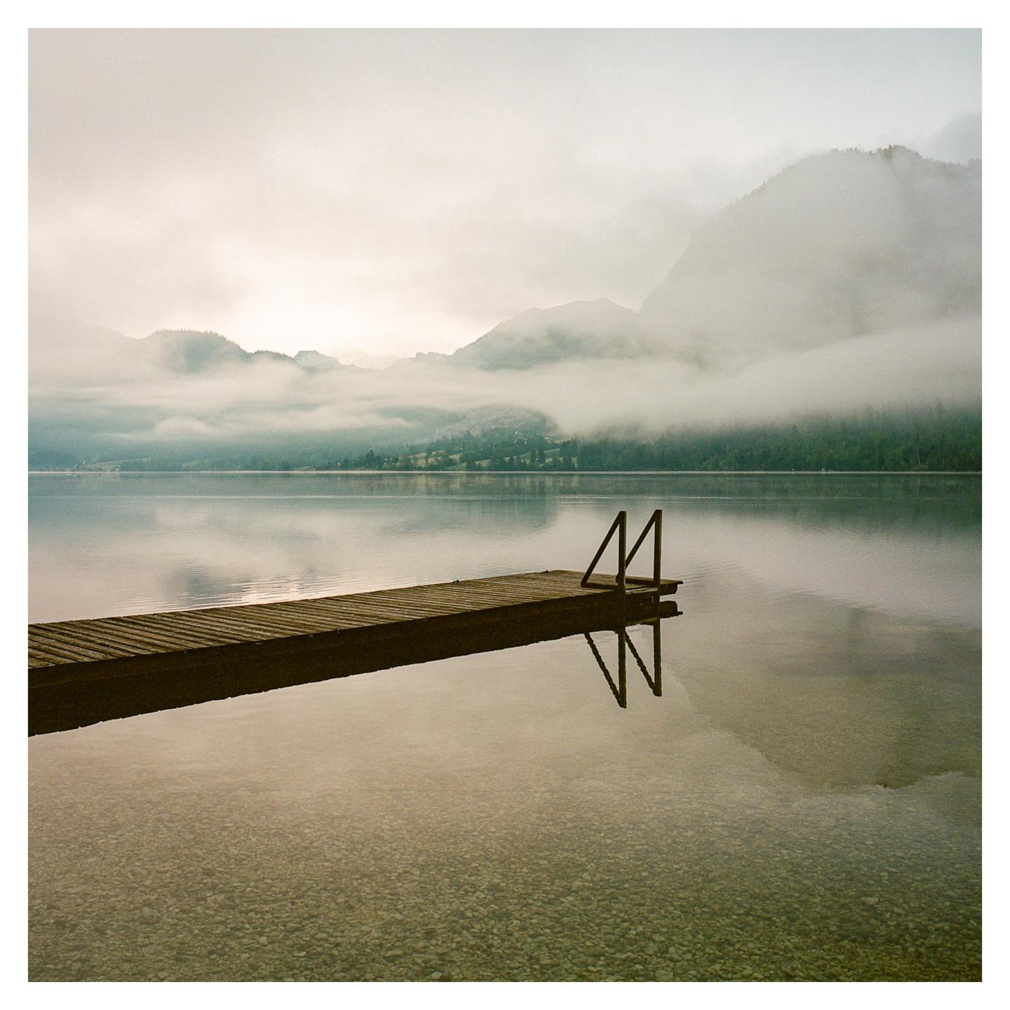 A wooden dock with a ladder extends into a calm lake surrounded by fog-covered mountains.