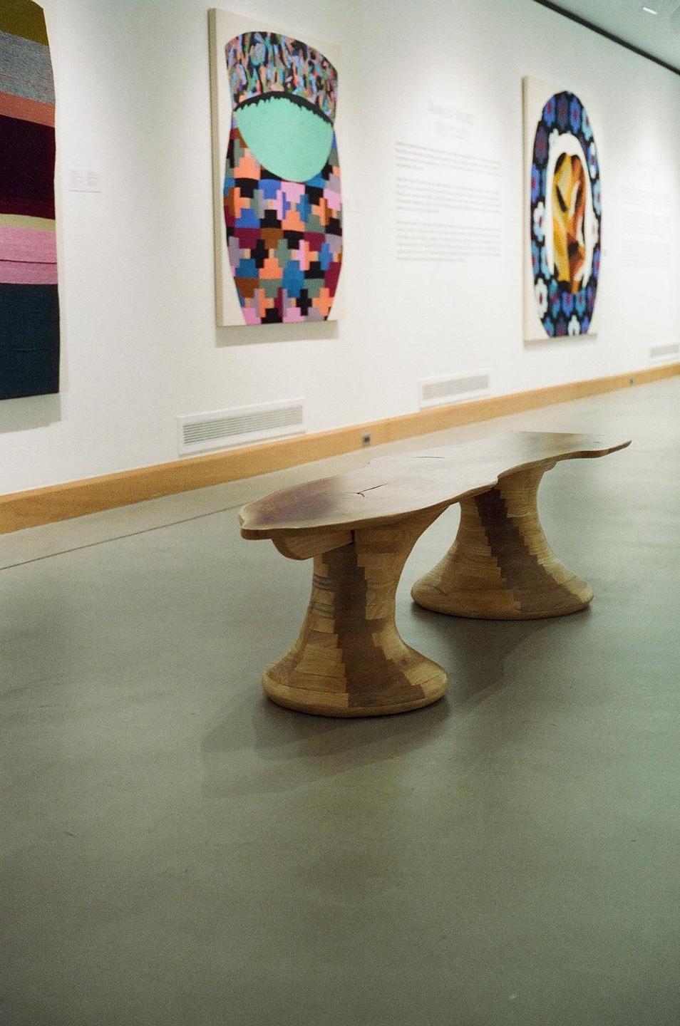A wooden bench with two bases sits on a gray floor in a gallery with colorful artworks on the wall.