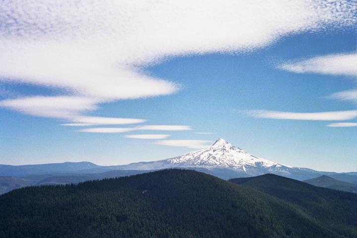 A snow-capped mountain peak rises above a forested mountain range under a blue sky with scattered clouds.