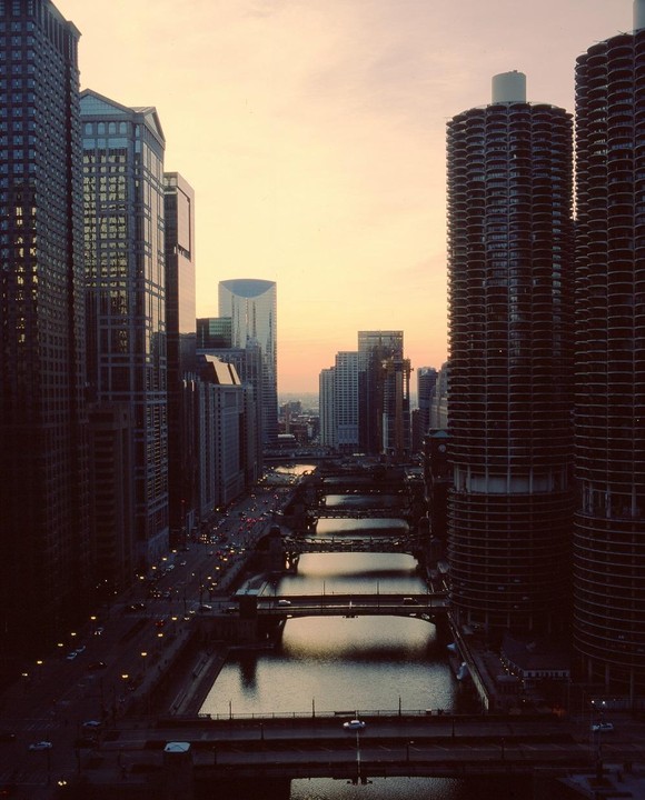 A high-angle view shows the Chicago River with multiple bridges, surrounded by tall buildings and city streets at dusk.