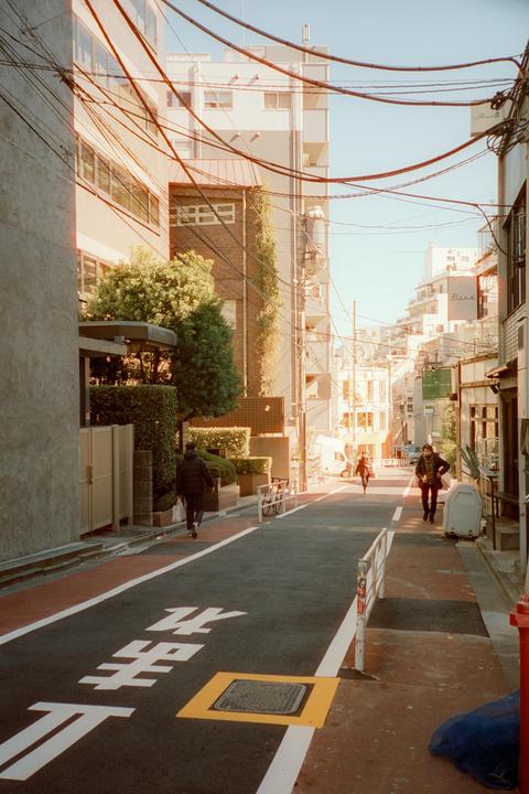 Three people walk along a narrow street lined with buildings and power lines under afternoon light.