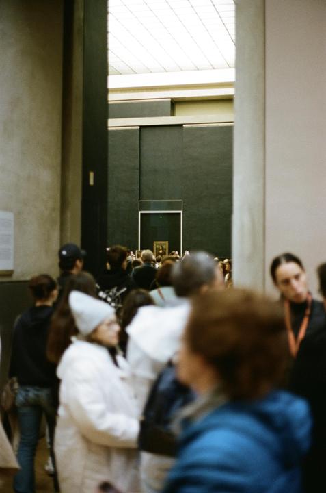 A crowd of people stands in a museum hall, looking at a painting in the distance.
