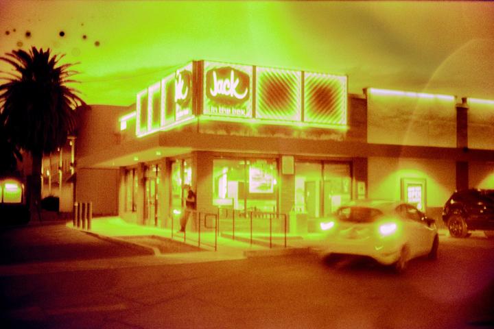 A Jack in the Box restaurant is brightly lit at night with a palm tree to its left and cars in front.