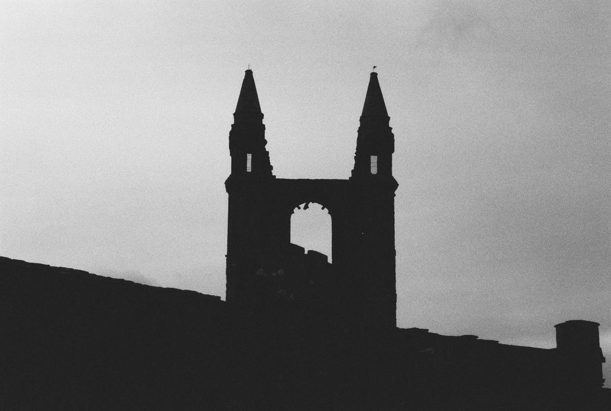 A silhouette of a ruined church tower with two spires stands against a cloudy sky.