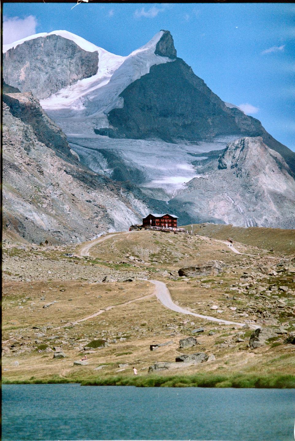 A red building sits on a hill overlooking a lake, with a snow-capped mountain in the background.