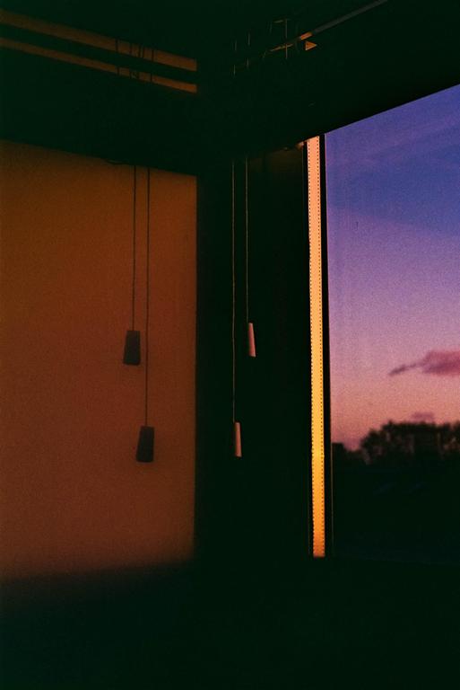 A window with blinds and pull cords looks out onto a colorful sunset sky with trees in the distance.