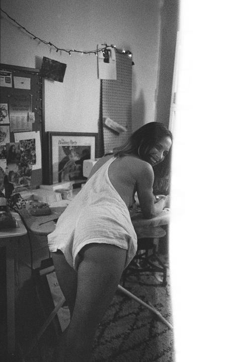 A woman in a white romper smiles at the camera while leaning on a desk in a room with string lights and a bulletin board.