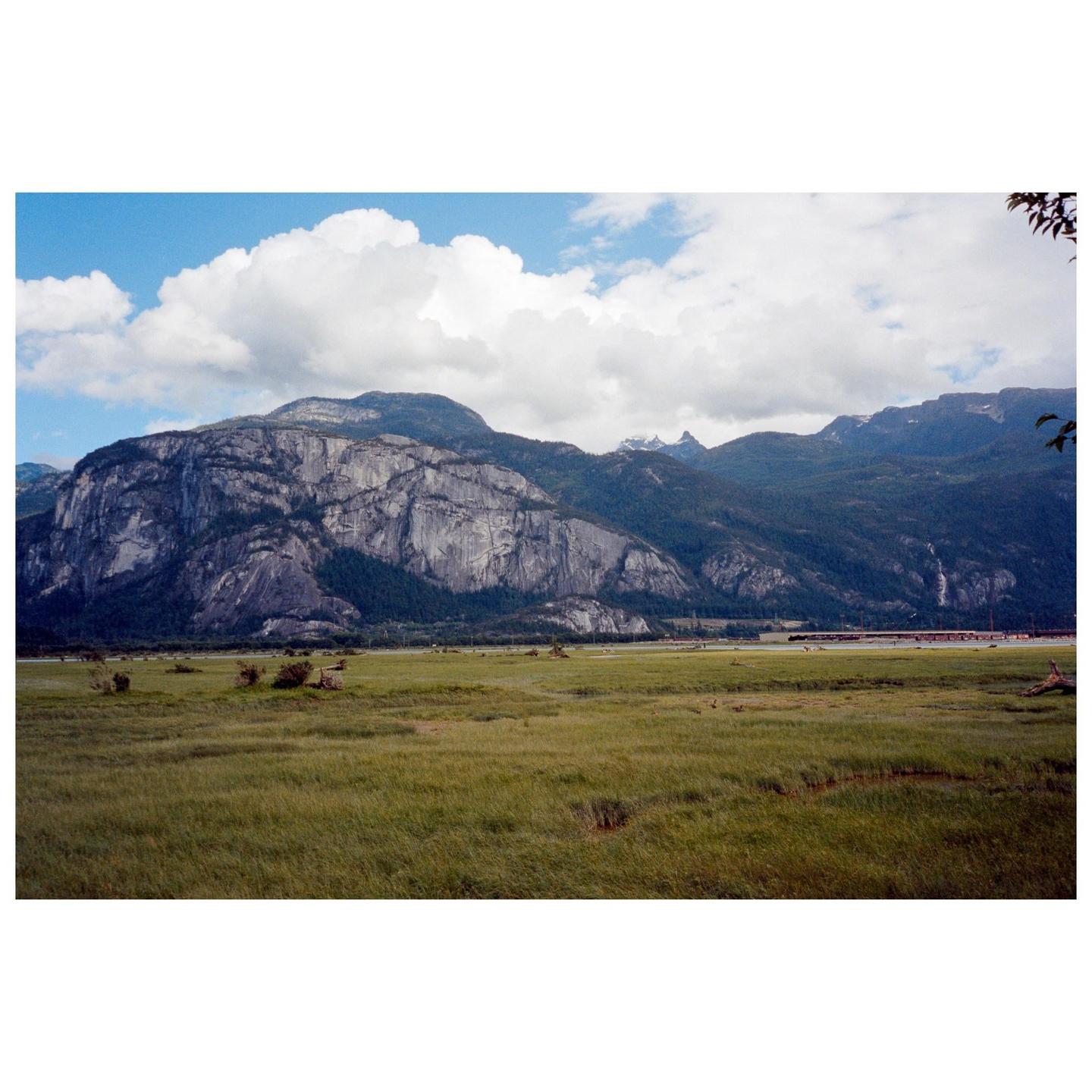A vast green field stretches towards a large mountain range under a partly cloudy sky.