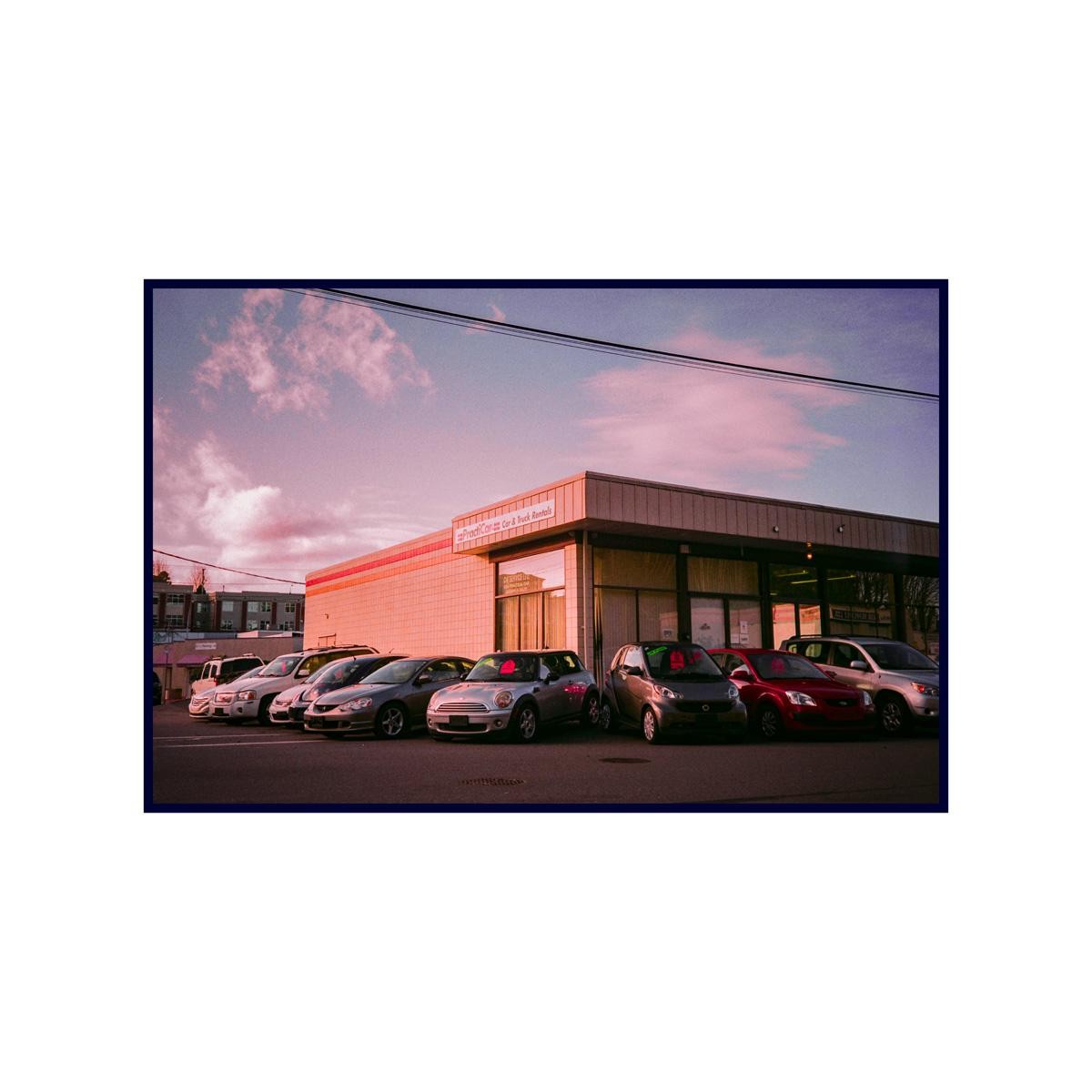 A car rental building with several cars parked in front of it is visible under a pink and purple sky.