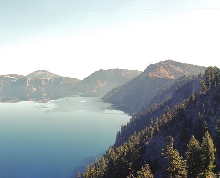 A wide view of Crater Lake shows the deep blue water surrounded by tree-covered mountains under a bright sky.