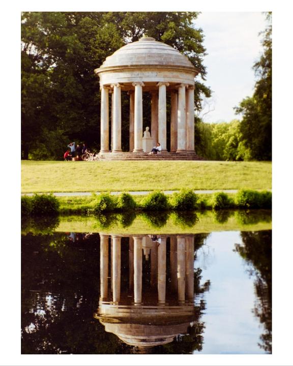 A classical rotunda with columns is reflected in a body of water, with people gathered on the grass nearby.